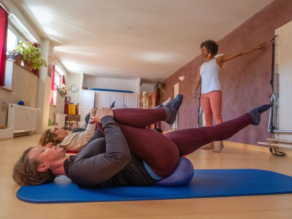 un groupe d'élèves suivent un cours collectif de Pilates avec petit ballon sous la direction d'une enseignante qui indique l'intention du mouvement par des gestes